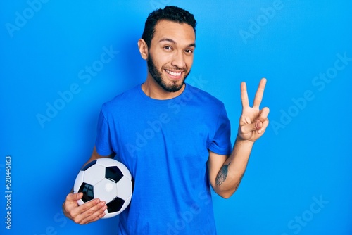 Hispanic man with beard holding soccer ball smiling looking to the camera showing fingers doing victory sign. number two. © Krakenimages.com