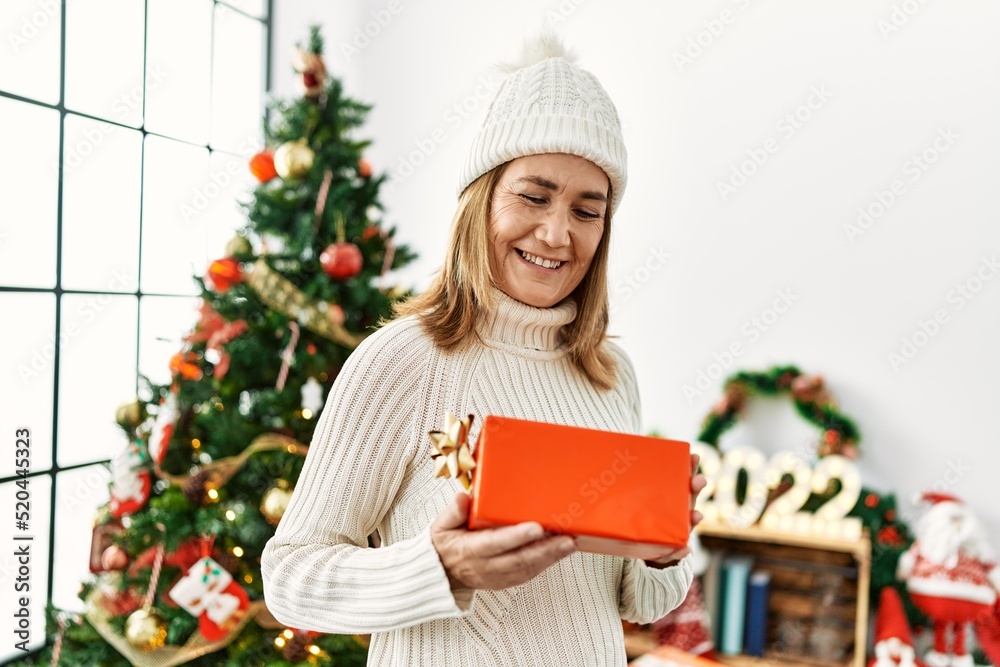 Middle age caucasian woman smiling confident holding gift standing by christmas tree at home Middle age caucasian woman smiling confident holding gift standing by christmas tree at home