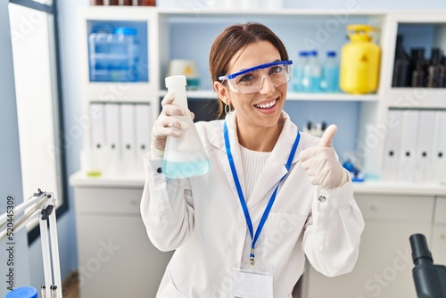 Young brunette woman working at scientist laboratory smiling happy and positive  thumb up doing excellent and approval sign