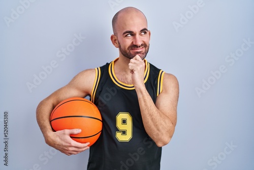 Young bald man with beard wearing basketball uniform holding ball with hand on chin thinking about question, pensive expression. smiling and thoughtful face. doubt concept.