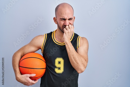 Young bald man with beard wearing basketball uniform holding ball looking stressed and nervous with hands on mouth biting nails. anxiety problem.