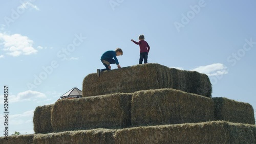 Two boys scale big pyramid pile of hay bales on autumn afternoon