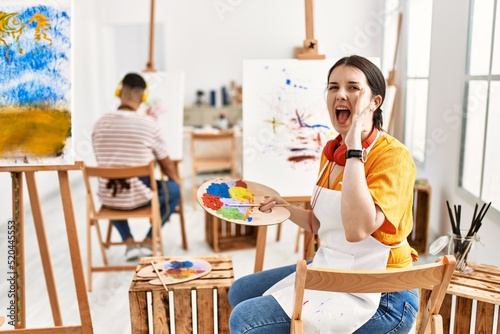 Young artist woman painting on canvas at art studio shouting and screaming loud to side with hand on mouth. communication concept. © Krakenimages.com