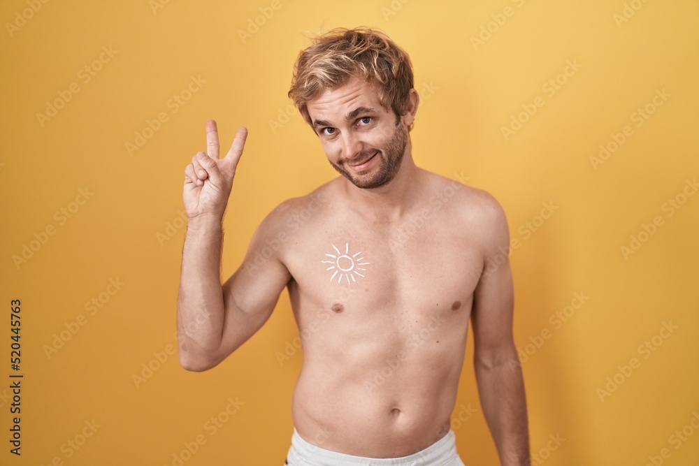 Caucasian man standing shirtless wearing sun screen smiling looking to the camera showing fingers doing victory sign. number two.