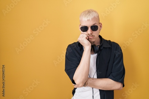 Young caucasian man wearing sunglasses standing over yellow background thinking looking tired and bored with depression problems with crossed arms.