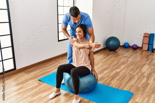 Latin man and woman wearing physiotherapist uniform having rehab session using fit ball at rehab center