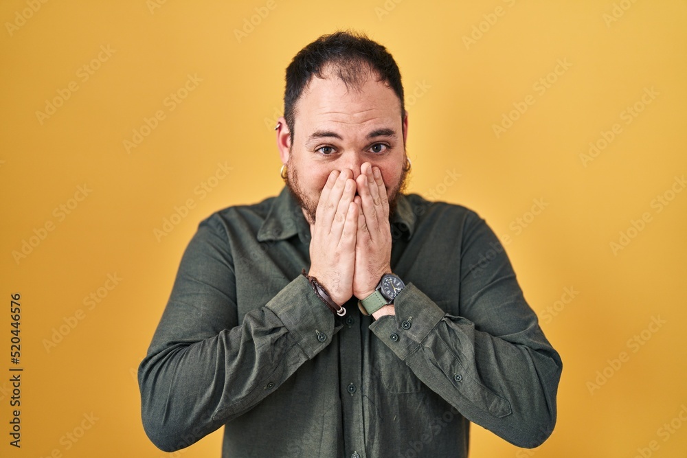 Plus size hispanic man with beard standing over yellow background laughing and embarrassed giggle covering mouth with hands, gossip and scandal concept Plus size hispanic man with beard standing over yellow background laughing and embarrassed giggle covering mouth with hands, gossip and scandal concept
