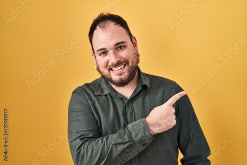 Plus size hispanic man with beard standing over yellow background cheerful with a smile on face pointing with hand and finger up to the side with happy and natural expression © Krakenimages.com