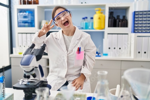 Hispanic girl with down syndrome working at scientist laboratory doing peace symbol with fingers over face  smiling cheerful showing victory