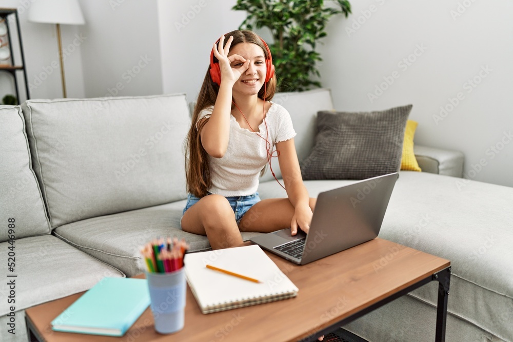 Young brunette teenager doing homework with laptop at home smiling happy doing ok sign with hand on eye looking through fingers Young brunette teenager doing homework with laptop at home smiling happy doing ok sign with hand on eye looking through fingers