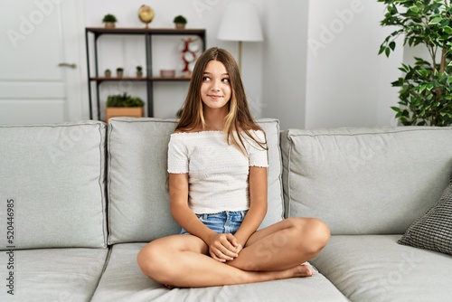 Young brunette teenager sitting on the sofa at home smiling looking to the side and staring away thinking.