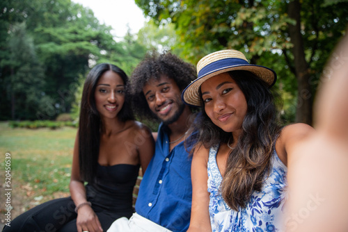 Portrait of three smiling friends sitting on a bench taking a selfie in a public park