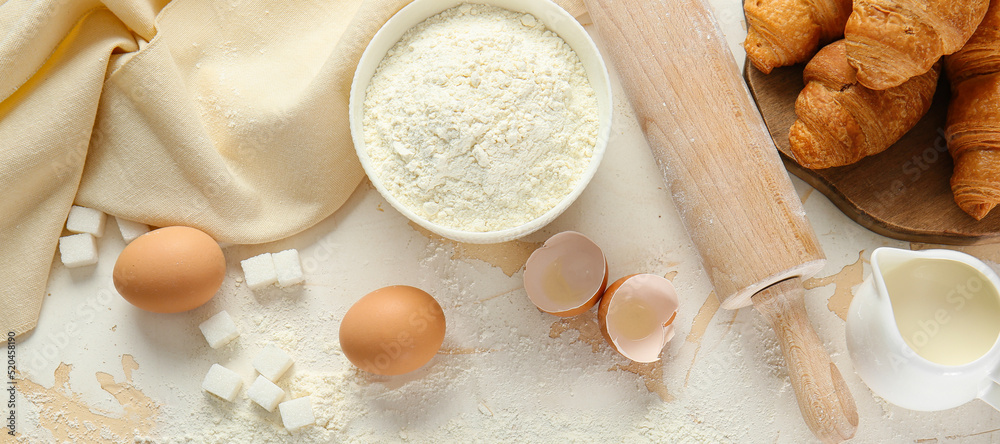 Bowl of wheat flour with raw eggs, sugar, milk, rolling pin and baked croissants on white table Bowl of wheat flour with raw eggs, sugar, milk, rolling pin and baked croissants on white table