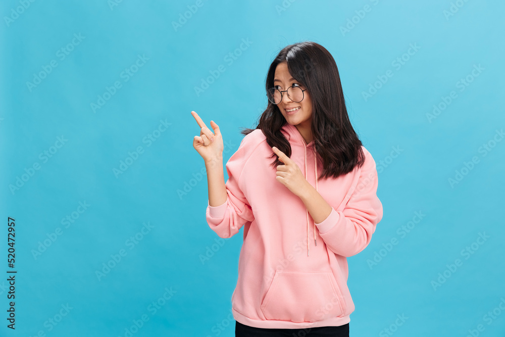 Cheerful Asian student young lady in pink hoodie sweatshirt wear round glasses point fingers aside posing isolated on over blue background. The best offer for ad. Eyewear for vision correction concept Cheerful Asian student young lady in pink hoodie sweatshirt wear round glasses point fingers aside posing isolated on over blue background. The best offer for ad. Eyewear for vision correction concept
