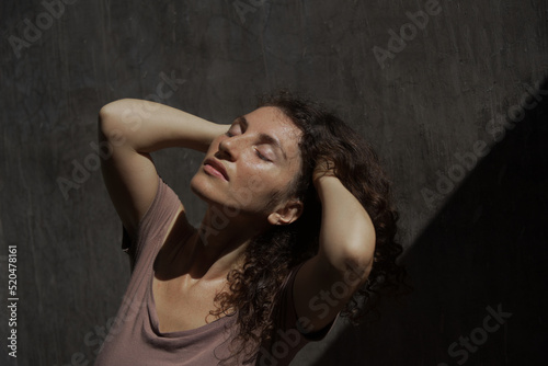 Psychological portrait of a curly woman in a dark dress, who stands with her eyes closed and her face up to the light. Concept of stress, depression, religion. Dark gray background with copy space.