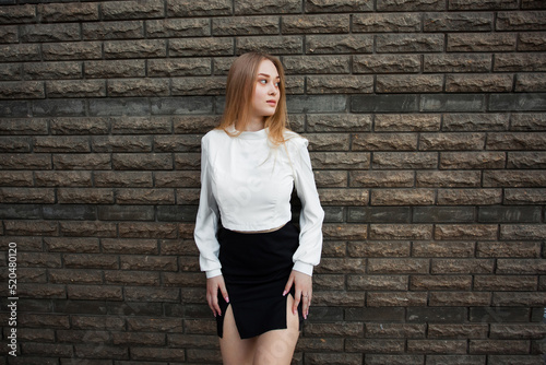 A slender young schoolgirl with blond hair, in a white shirt and a short black skirt, stands against a dark textured stone wall, looks away and keeps her hands on her hips. photo