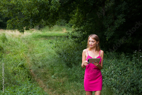 A slender young freelancer girl with long blond hair  in a pink dress  stands in the forest on a green lawn and remotely works and studies on a tablet.