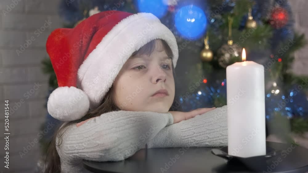 Child look at candle in decorated room. An upset little girl sits on the table with burning candle in the New Year room. Child look at candle in decorated room. An upset little girl sits on the table with burning candle in the New Year room.