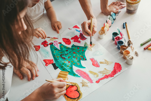 children with their mother draw a drawing with paints and brushes, gouache, an elegant Christmas tree, a children's drawing on the table