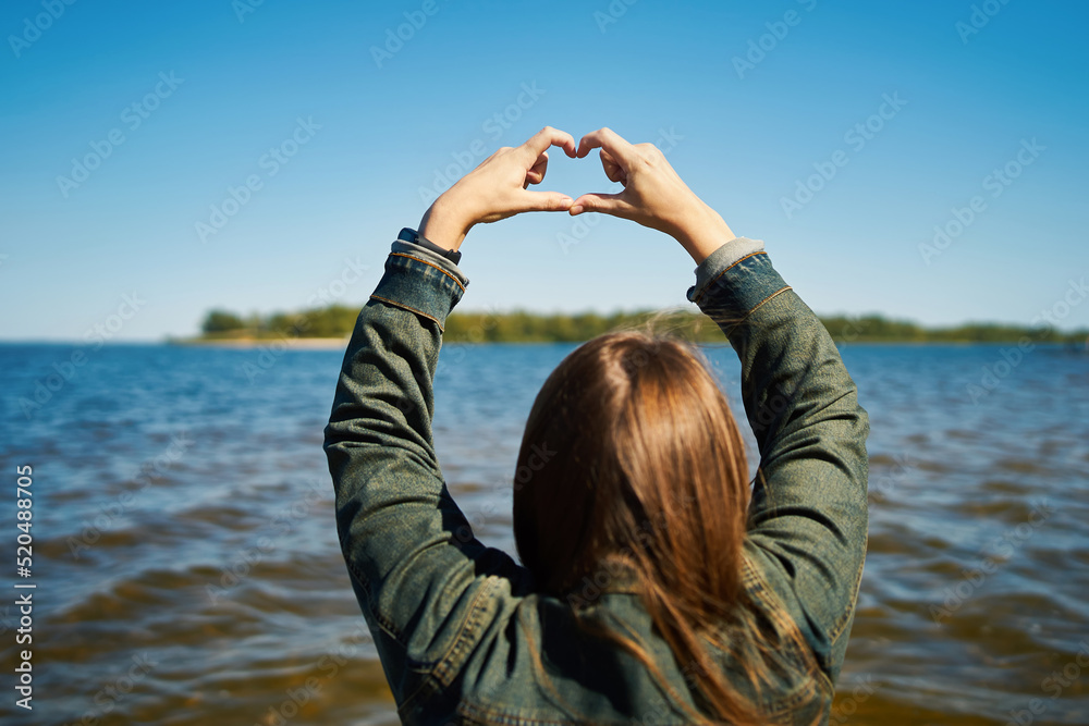 The girl makes a heart with her hands against the background of a saturated blue sky and a river, rear view