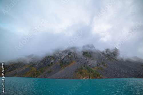 Dramatic tranquil meditate landscape with ripples on big turquoise mountain lake against high sharp rocks in low clouds. Calm azure alpine lake and large rocky mountain range in low cloudy gray sky.