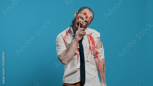Creepy dead walking corpse making shush secrecy hand gesture on blue background. Dangerous walking dead corpse with deep and bloody wounds being confidential while smirking at camera.