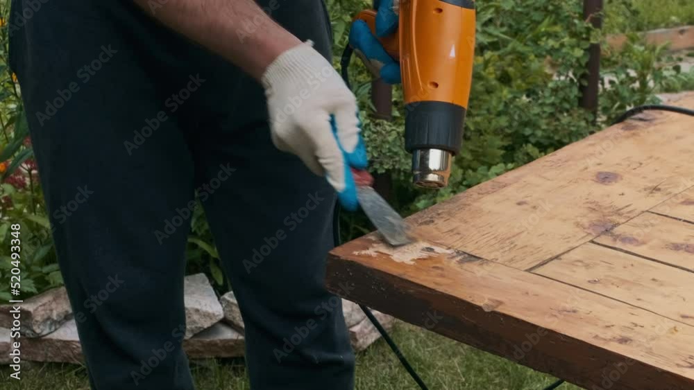 Man removing old varnish and paint from wood using scraper and heat gun. Restoration work of old wooden table outdoors. Furniture repair, reuse of old things concept. Man removing old varnish and paint from wood using scraper and heat gun. Restoration work of old wooden table outdoors. Furniture repair, reuse of old things concept.