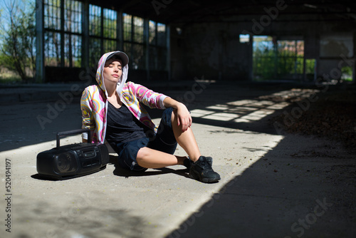 Young woman street dancer sitting in abandoned building and listening to music from cassette player on a sunny summer day