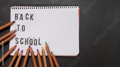 blank page of a notebook with the phrase "Back To School" written in English with worn up letters next to colored pencils on a black chalkboard background