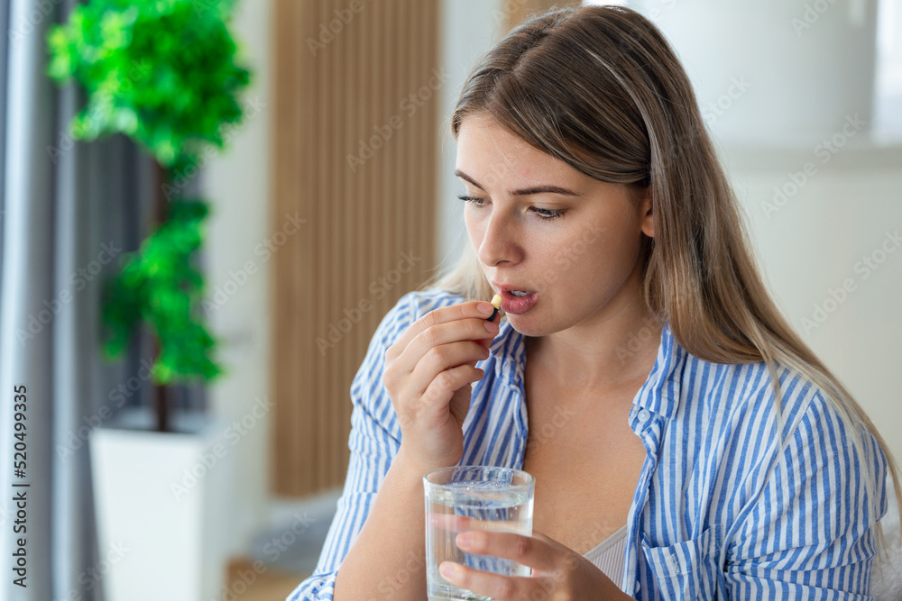 Concept of suffering from terrible pain. Beautiful, sad, upset, unhappy, troubled, weak woman wearing casual clothes is sitting on a sofa and taking a pill Concept of suffering from terrible pain. Beautiful, sad, upset, unhappy, troubled, weak woman wearing casual clothes is sitting on a sofa and taking a pill