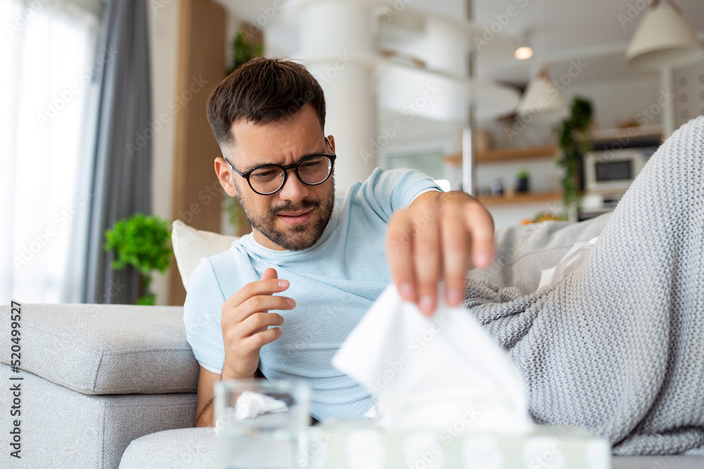 Sick sad man with glasses sits on couch at home suffers from runny nose flu disease coronavirus pandemic covid epidemic sneezes. Unwell guy feeling bad fever virus illness symptoms indoor