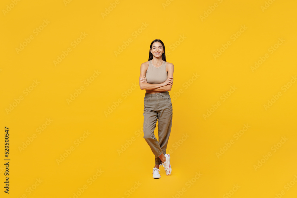 Full body smiling happy fun young latin woman 30s she wear basic beige tank shirt looking camera hold hands crossed folded isolated on plain yellow backround studio portrait People lifestyle concept