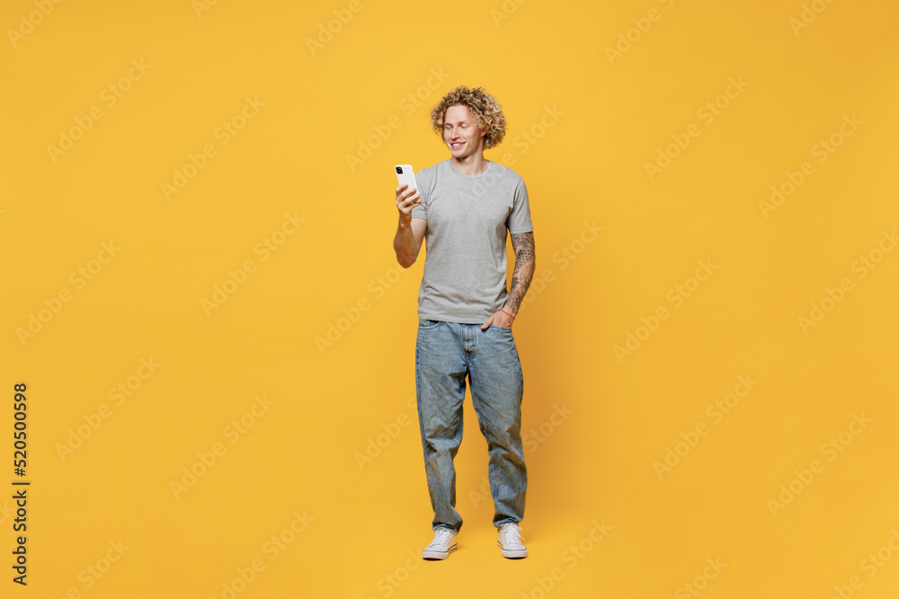Fototapeta premium Full body young smiling cheerful fun satisfied caucasian man 20s he wear grey t-shirt hold in hand use mobile cell phone isolated on plain yellow backround studio portrait. People lifestyle concept.