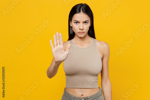 Young strict sad latin woman 30s she wearing basic beige tank shirt showing stop gesture with palm refusing reject say no isolated on plain yellow backround studio portrait. People lifestyle concept.