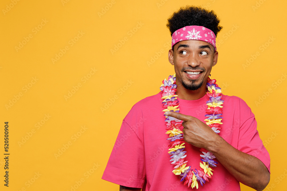 Young smiling man 20s he wear pink t-shirt hawaiian lei bandana near hotel pool point index finger aside on workspace area mock up isolated on plain yellow background Summer vacation sea rest concept