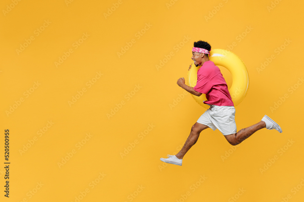 Side view happy fun cool young man he wear pink t-shirt near hotel pool jump high run fast hurrying up hold inflatable rubber ring isolated on plain yellow background Summer vacation sea rest concept