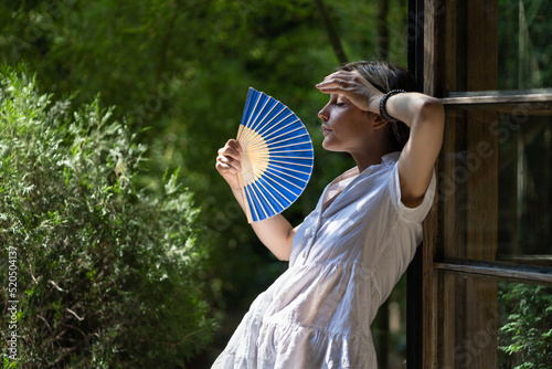 Young woman suffering heatstroke outdoors using fan to get fresh air. Unhappy girl feel bad of hot temperature outside. Frustrated female tired of summer season heat in fatigue touch forehead sweating photo