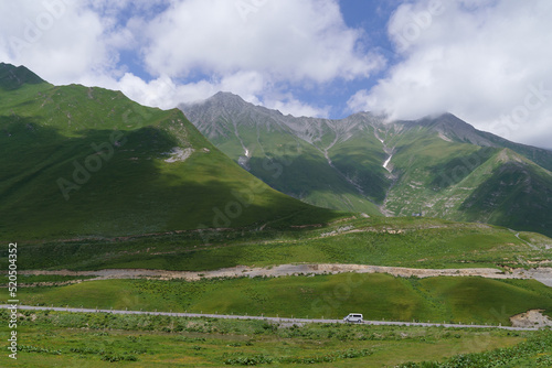 Cross pass and car driving on Georgian military road in highland on sunny summer day under blue sky with white cumulus clouds. Giant mountains covered with alpine meadows and forests in Georgia