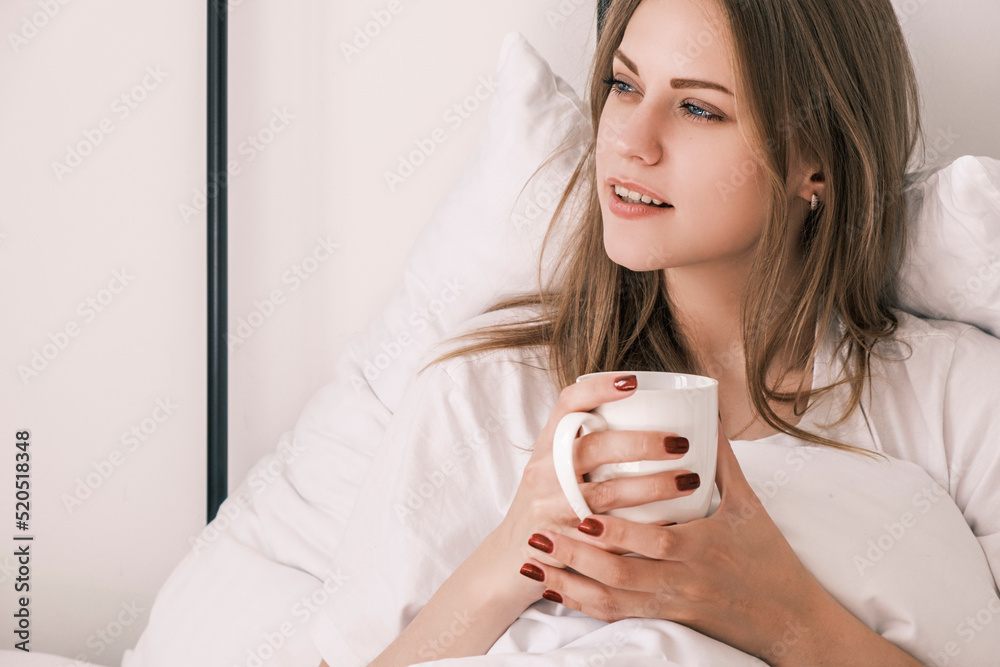 Young attractive woman lies in bed after waking up with white cup in her hand looking out the window. Morning routine, time for yourself. Contemplation and enjoyment. Psychological self care