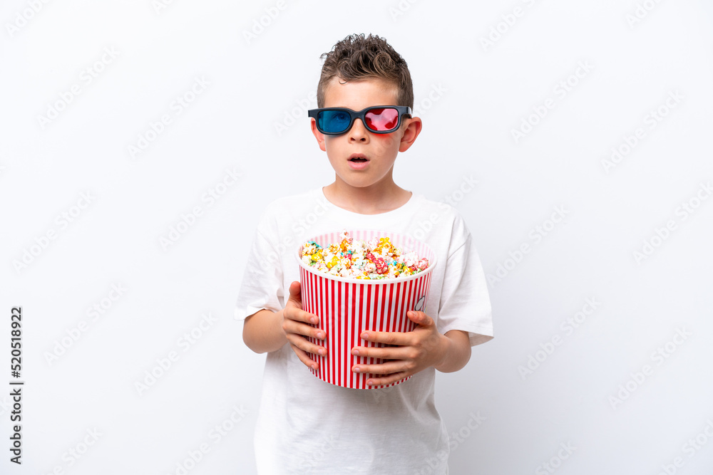 Little caucasian boy isolated on white background surprised with 3d glasses and holding a big bucket of popcorns