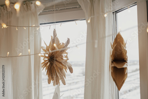Stylish paper christmas stars and lights hanging in window in festive decorated boho room. Handmade paper swedish stars and garland, scandinavian decoration. Atmospheric christmas time