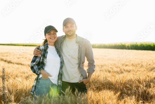 A couple of farmers in plaid shirts and caps stand embracing on agricultural field of wheat at sunset © scharfsinn86