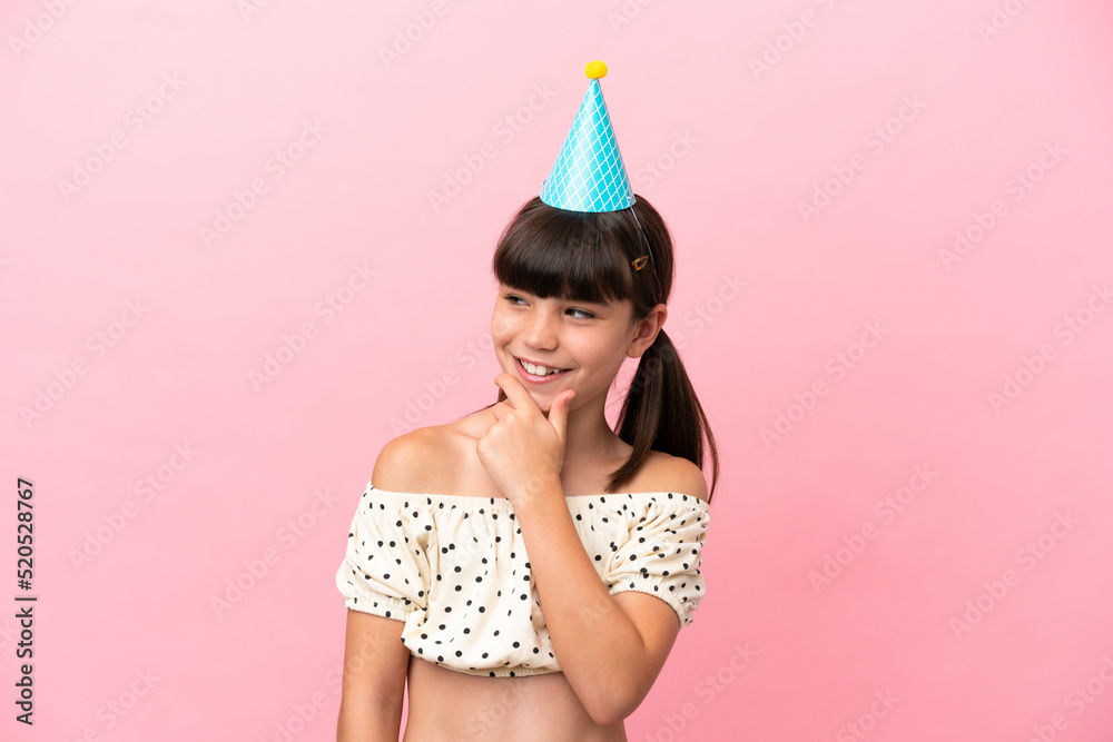 Little caucasian kid with birthday hat isolated on pink background looking to the side and smiling