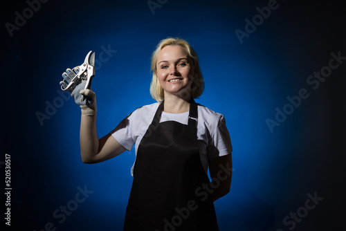 A blond gardener woman in a black apron and protective glove holds garden shears in her hand. Blue background.
