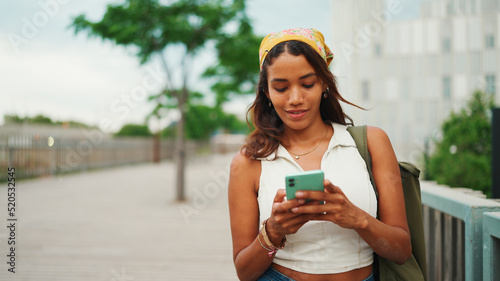 Cute tanned woman with long brown hair wearing white top and yellow bandana walks on bridge with backpack on her shoulder and cell phone in her hand. Girl using phone photo