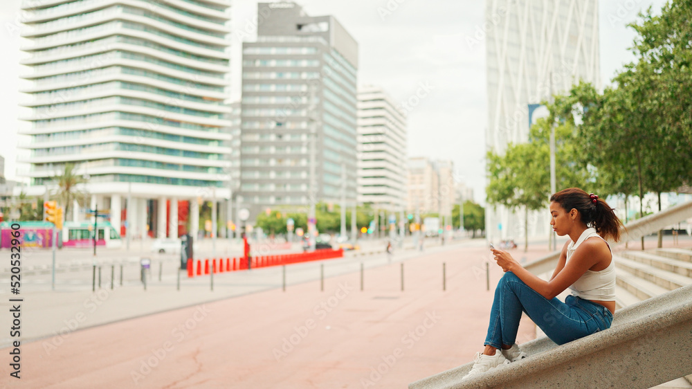 Profile of cute tanned woman with long brown hair wearing white top is using smartphone on city landscape background Profile of cute tanned woman with long brown hair wearing white top is using smartphone on city landscape background