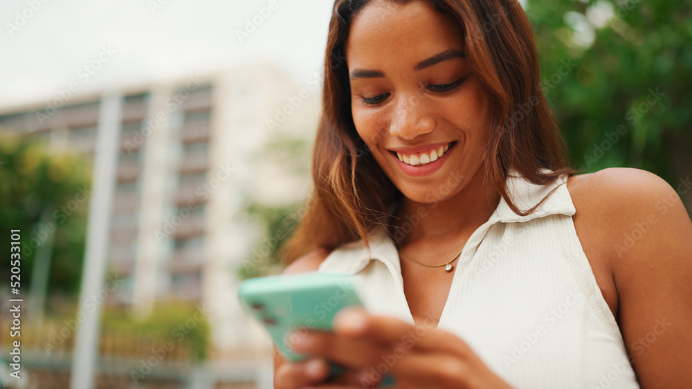 Clouse-up, beautiful girl with long dark hair wearing white top sits on bench and uses mobile phone. Young smiling woman writing in social networks on cellphone. Clouse-up, beautiful girl with long dark hair wearing white top sits on bench and uses mobile phone. Young smiling woman writing in social networks on cellphone.