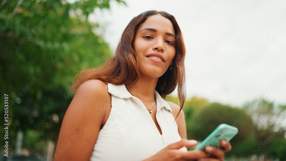 Clouse-up, beautiful girl with long dark hair wearing white top sits on bench and uses mobile phone. Young smiling woman writing in social networks on cellphone. Clouse-up, beautiful girl with long dark hair wearing white top sits on bench and uses mobile phone. Young smiling woman writing in social networks on cellphone.
