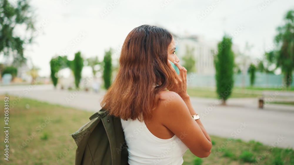 Beautiful girl with long dark hair wearing white top walks along path in city park and uses mobile phone. Young woman talking on cellphone