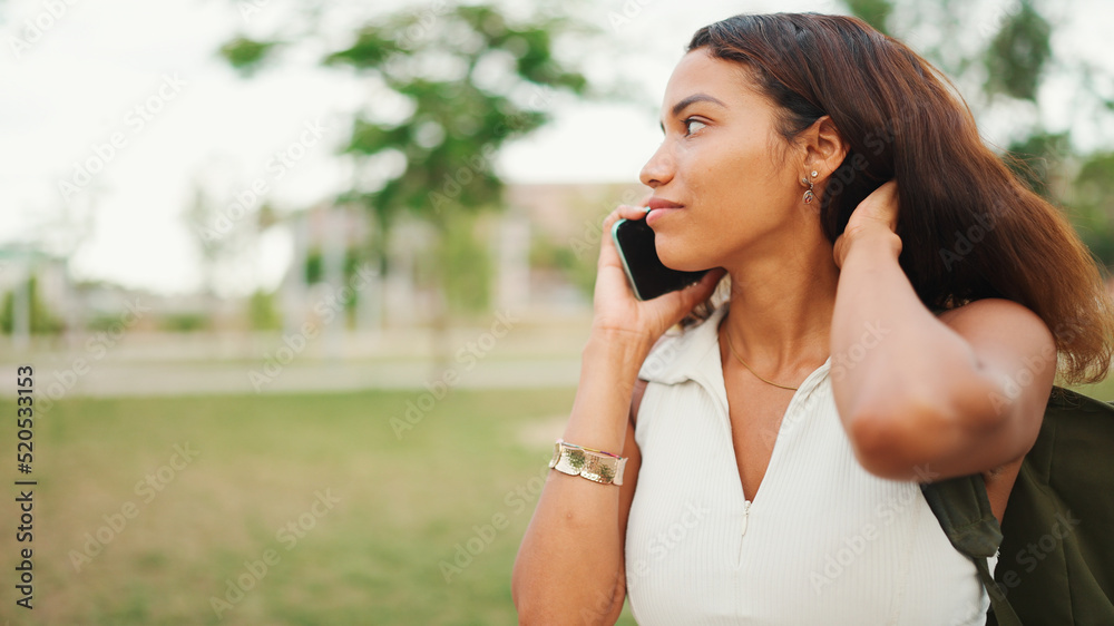 Beautiful girl with long dark hair wearing white top sits on bench in city park and uses mobile phone. Young woman talking on cellphone Beautiful girl with long dark hair wearing white top sits on bench in city park and uses mobile phone. Young woman talking on cellphone